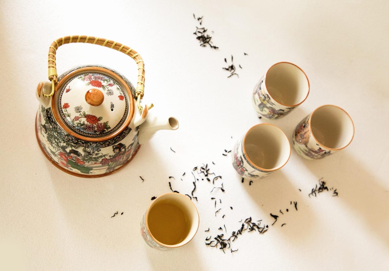 A teapot and small cups with loose leaf tea scattered on a light surface