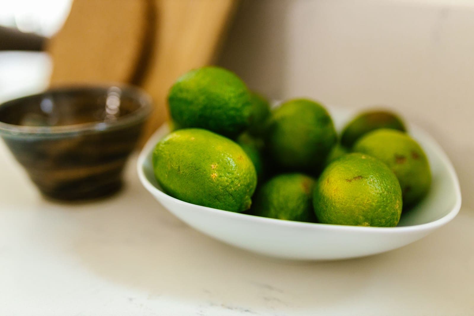 A close-up of fresh limes in a white bowl on a bright kitchen counter